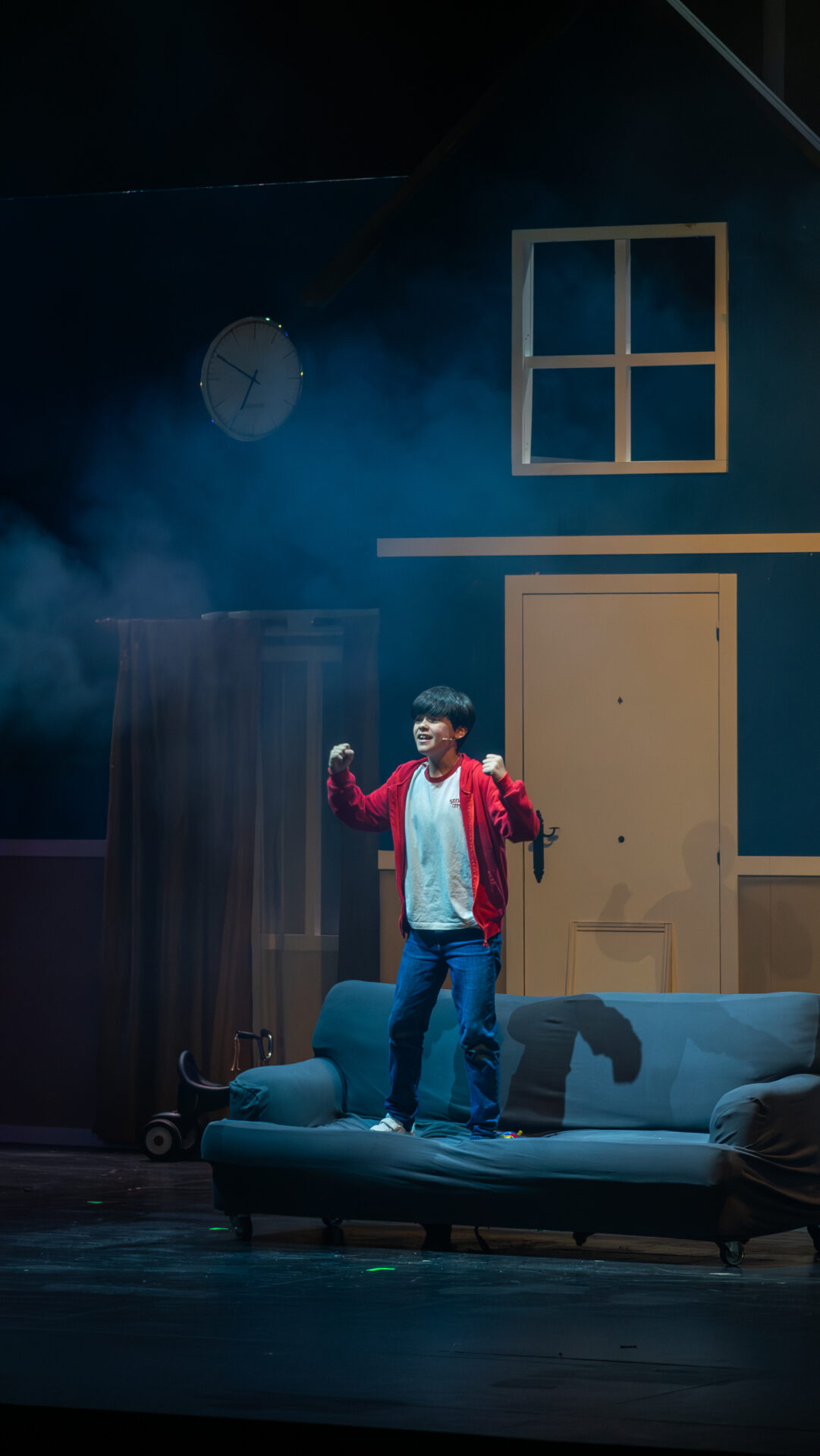 Escena del musical “Estoy Solo en Casa” con un personaje sobre un sofá, ambientado en una casa iluminada en azul. El musical familiar que no te puedes perder.